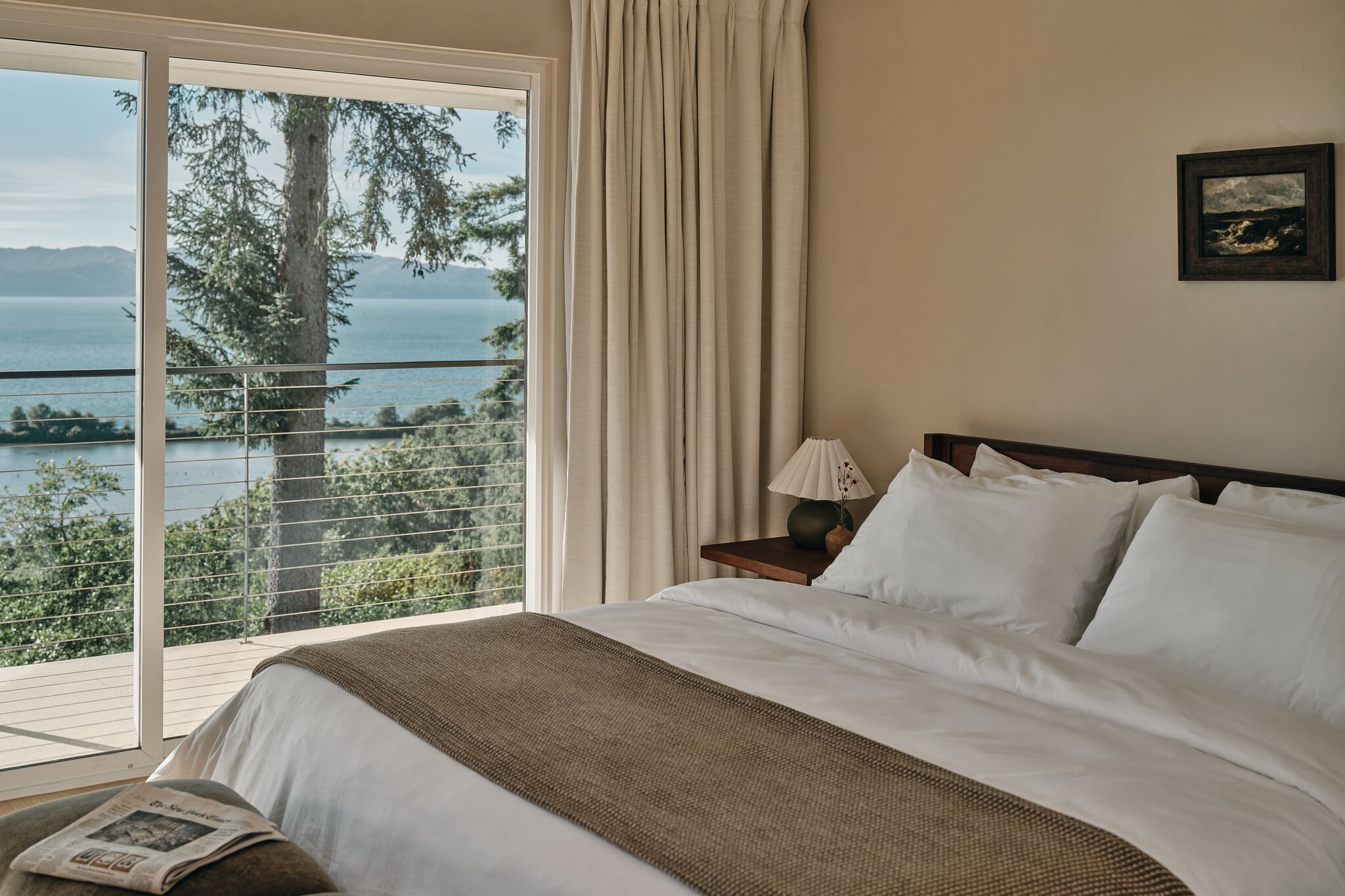 Ein Hotelzimmer mit einem großen Bett, einem Nachttisch, einer Lampe und Blick auf das Meer und Bäume durch eine Glasschiebetür.