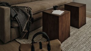 Una sala de estar moderna con un sofá, dos mesitas de madera, una mesa de centro, un bolso marrón y un cuadro en la pared.