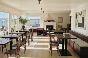 A spacious dining area with multiple tables and chairs, a fireplace, and a large window offering a view of the outdoors. - The Herondale Hotel (Astoria)