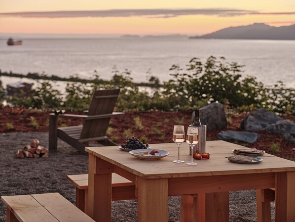 A wooden table set with wine glasses, a bottle, and plates of food on a beach with a chair and a pile of wood. - The Herondale Hotel (Astoria)