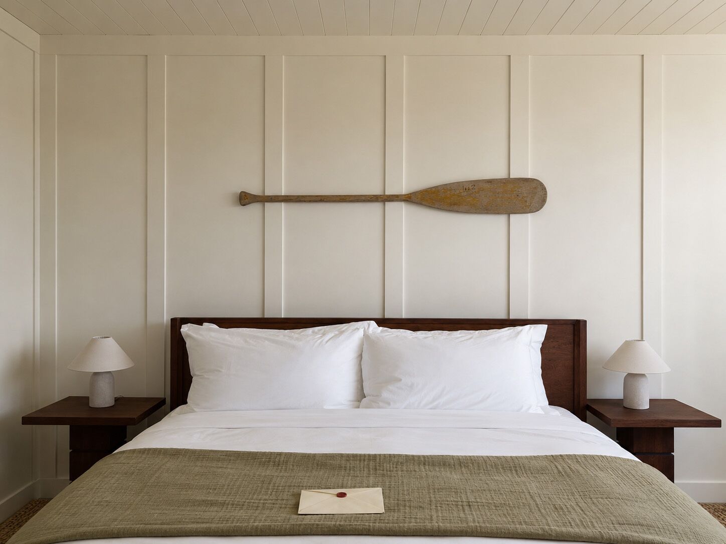 Una cama con sábanas blancas, dos mesitas de noche con lámparas, un cabecero de madera y una pala en la pared.