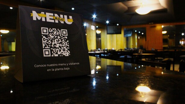 Placa de menu com código QR num restaurante, com mesas e cadeiras ao fundo.