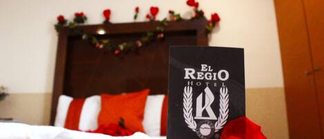 Habitación de hotel con una cama, un letrero que indica El Regio Hotel y pétalos de rosa roja sobre la cama.