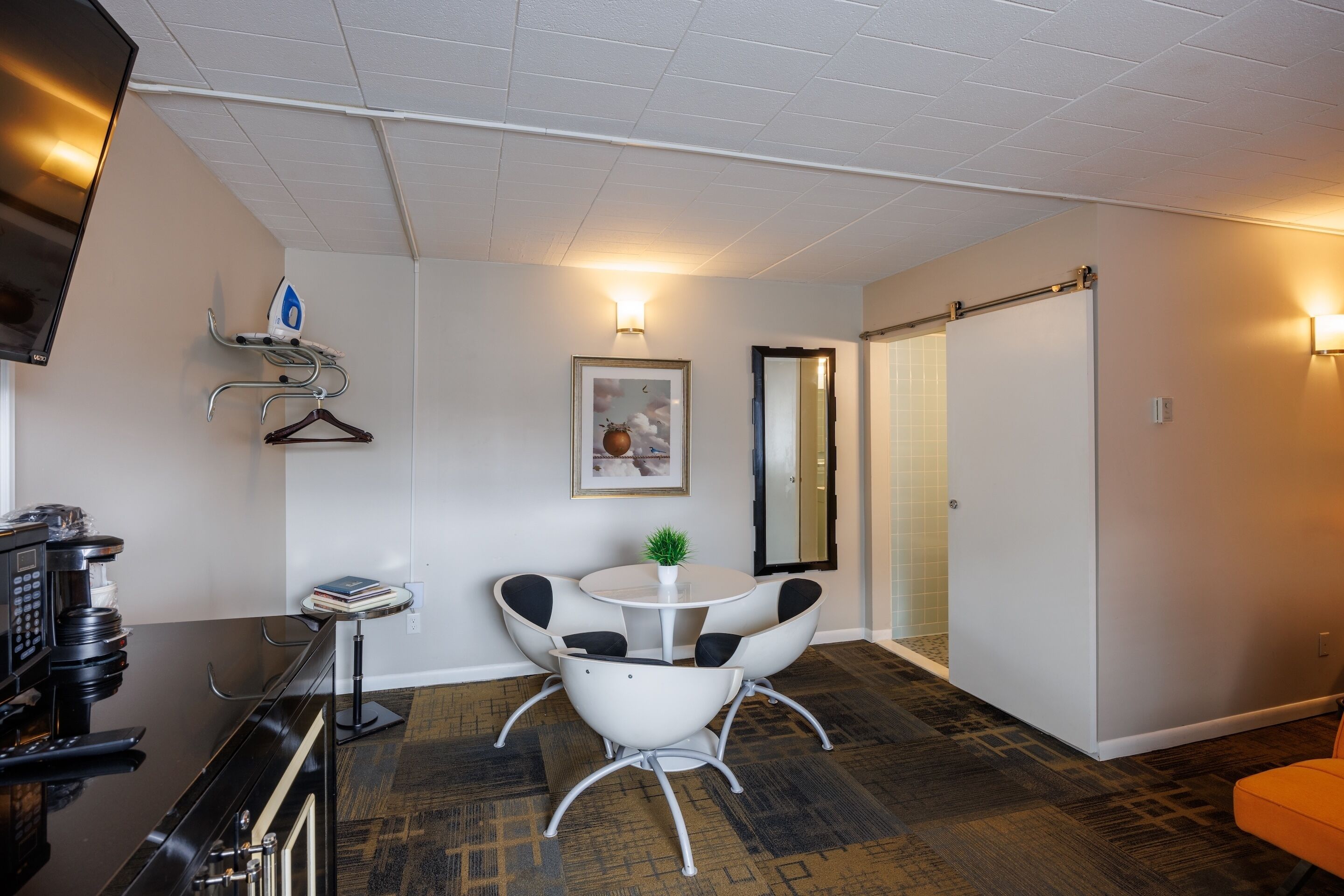Habitación de hotel con comedor que incluye una mesa y sillas blancas, una cafetera, un televisor y un perchero con colgadores.