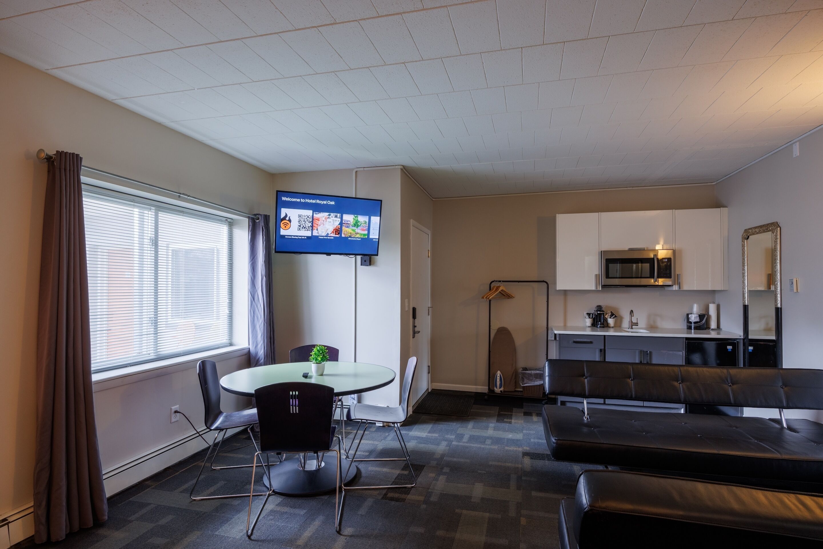 A modern hotel room with a round dining table, black chairs, a flat-screen TV, a kitchenette, and a seating area with a sofa and a bench.