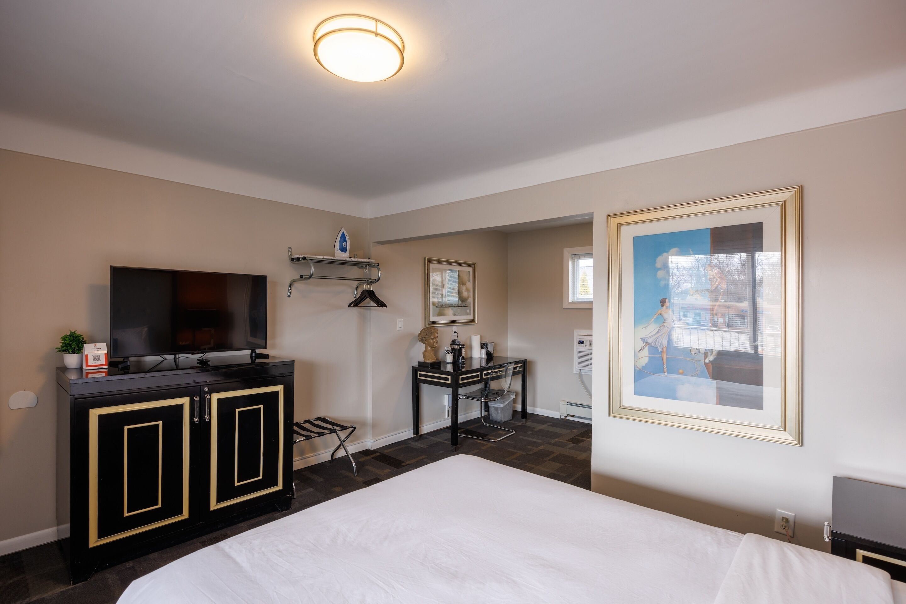 A hotel room with a bed, a desk with a chair, a television, a framed picture on the wall, and a small plant.
