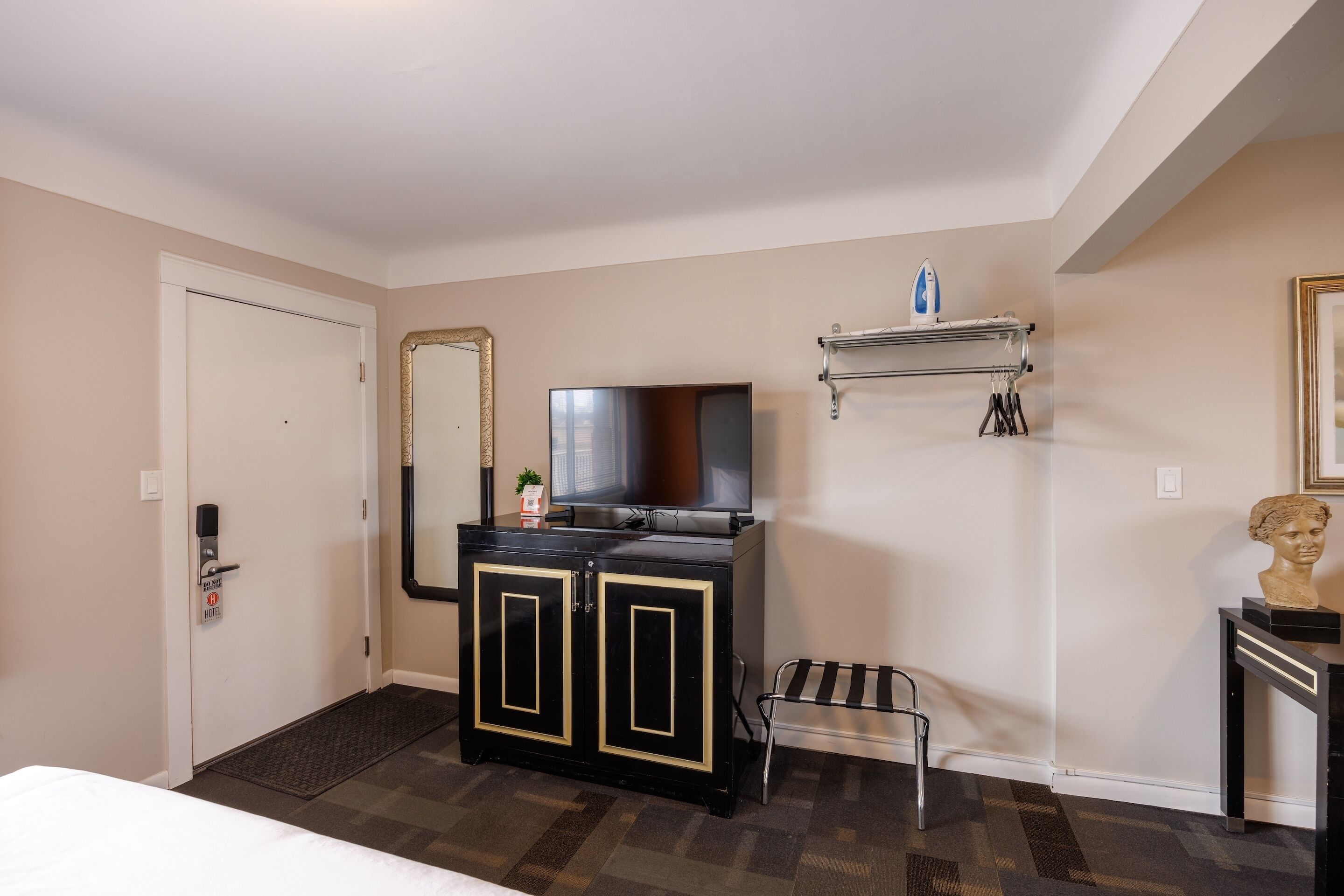 A hotel room with a bed, a black minibar, a television, a mirror, a small table with a bust, and a shoe rack.