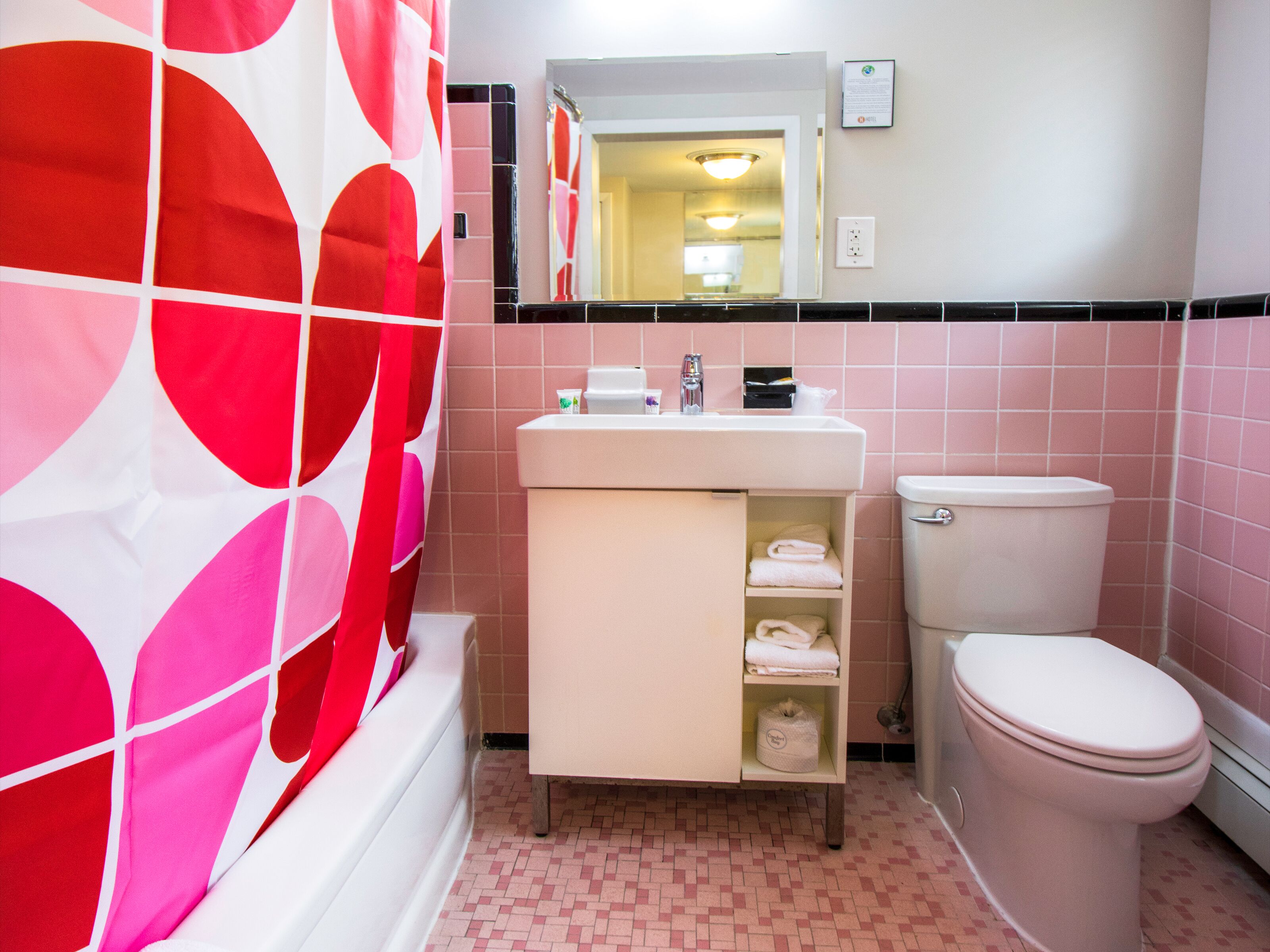 Une salle de bain avec un mur recouvert de carreaux roses et blancs, un lavabo blanc avec un miroir au-dessus, et des toilettes blanches.