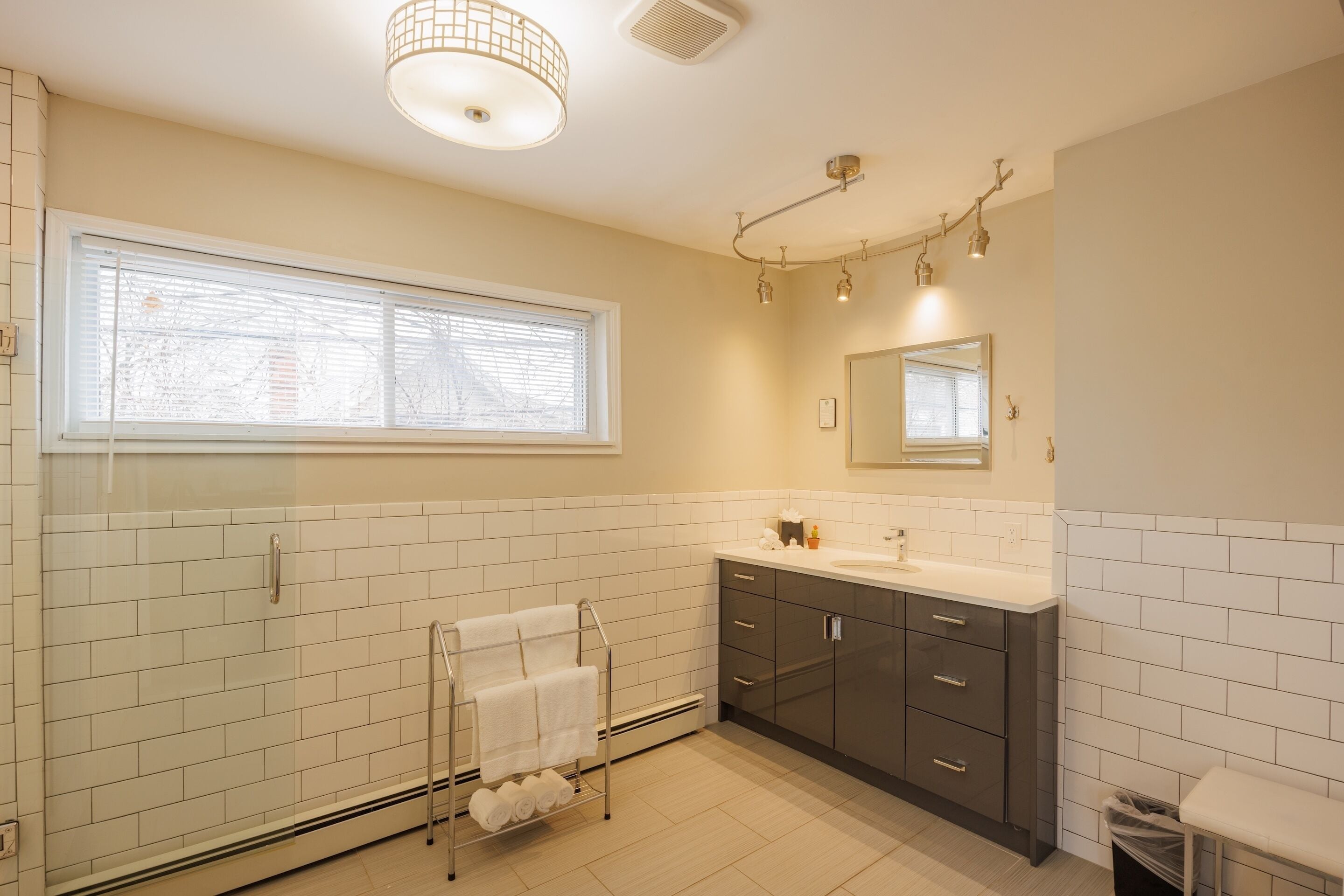A modern bathroom with white tiled walls, a large window, a vanity with dark cabinets, and a glass shower enclosure.