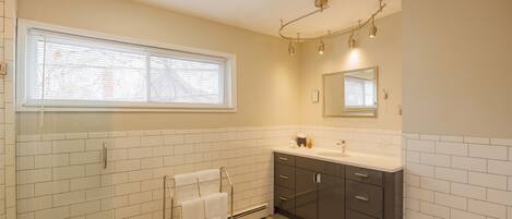 A modern bathroom with white tiled walls, a large window, a vanity with dark cabinets, and a glass shower enclosure.