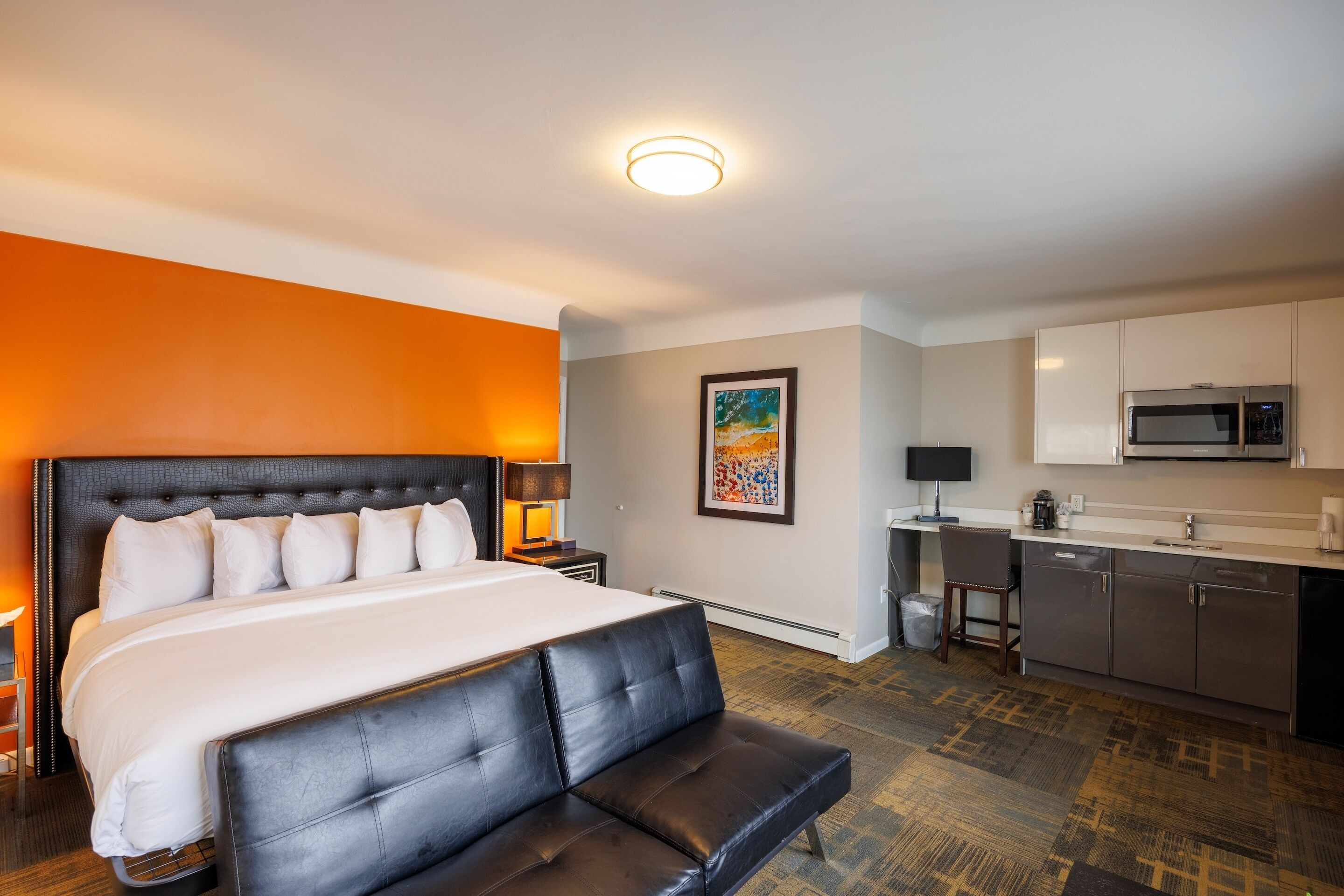 A hotel room with a large bed, a black leather bench, a kitchenette, and a painting on the wall.