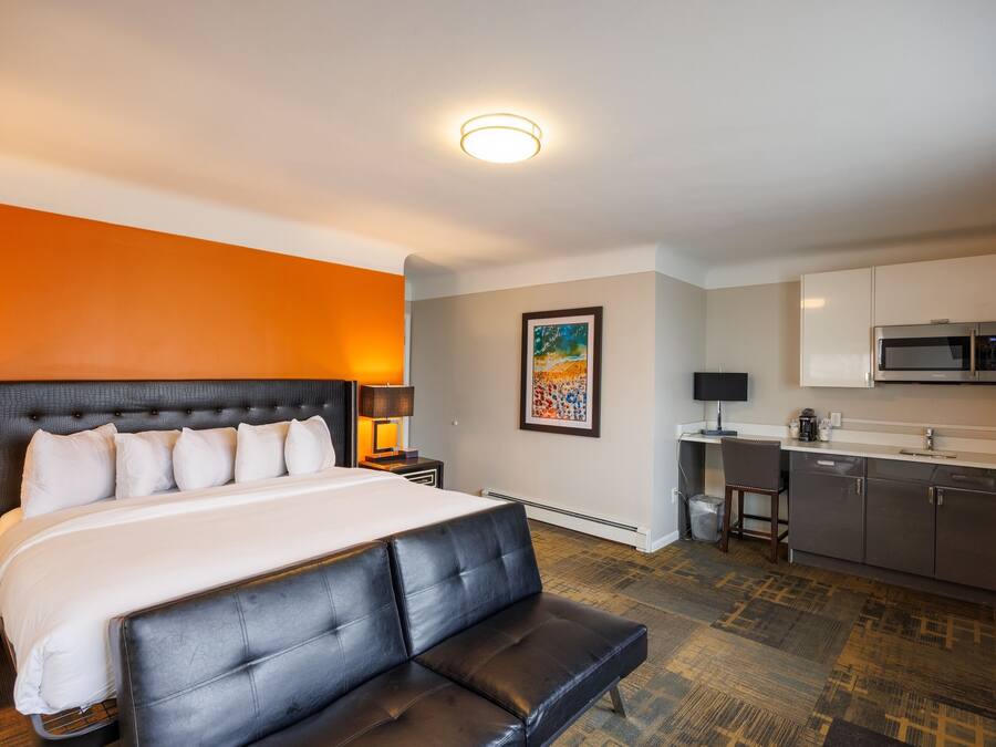 Quarto de hotel com uma cama grande, um banco de couro preto, uma kitchenette e uma pintura na parede.