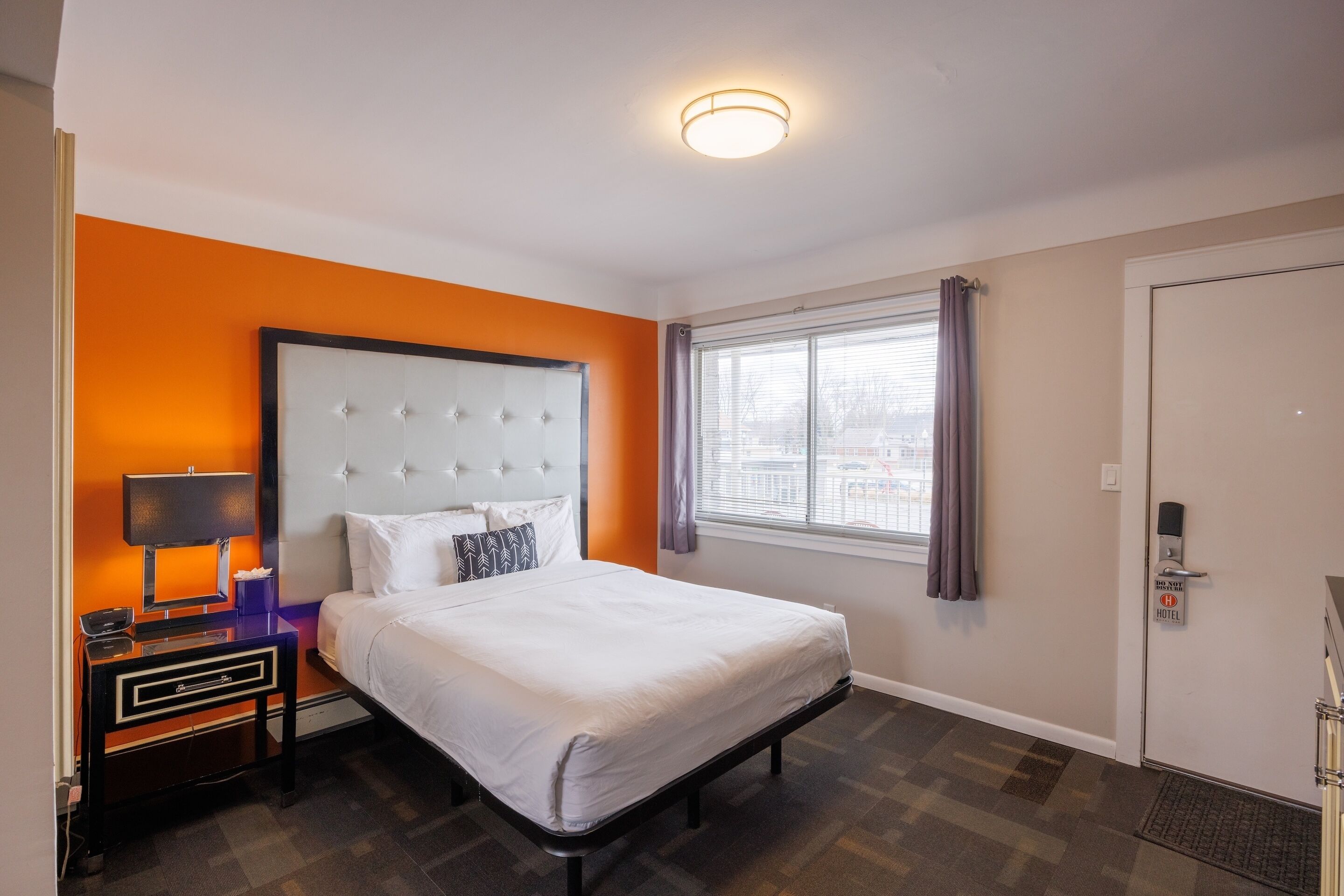 A hotel room with a bed, a nightstand with a lamp, a window with curtains, and a door with a hotel sign.