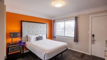 A hotel room with a bed, a nightstand with a lamp, a window with curtains, and a door with a hotel sign.