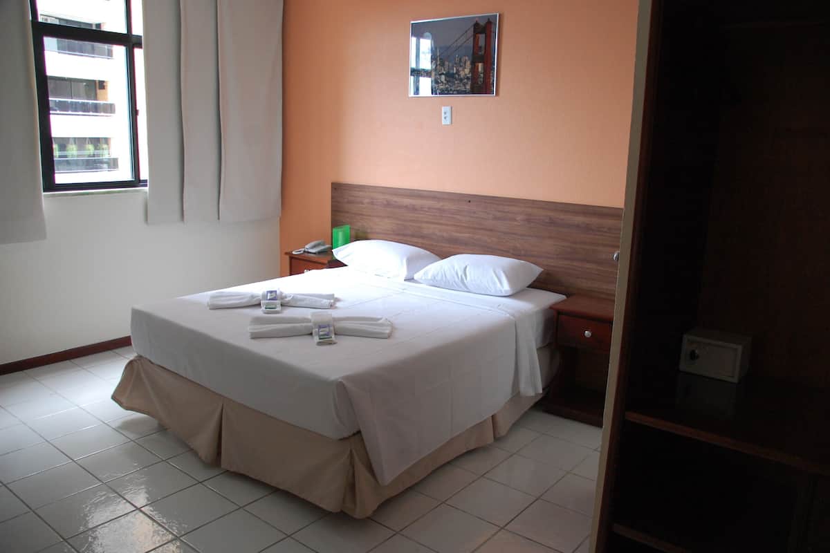 A neatly made bed with white linens and a wooden headboard, a bedside table with a phone and a green object, and a window with curtains.