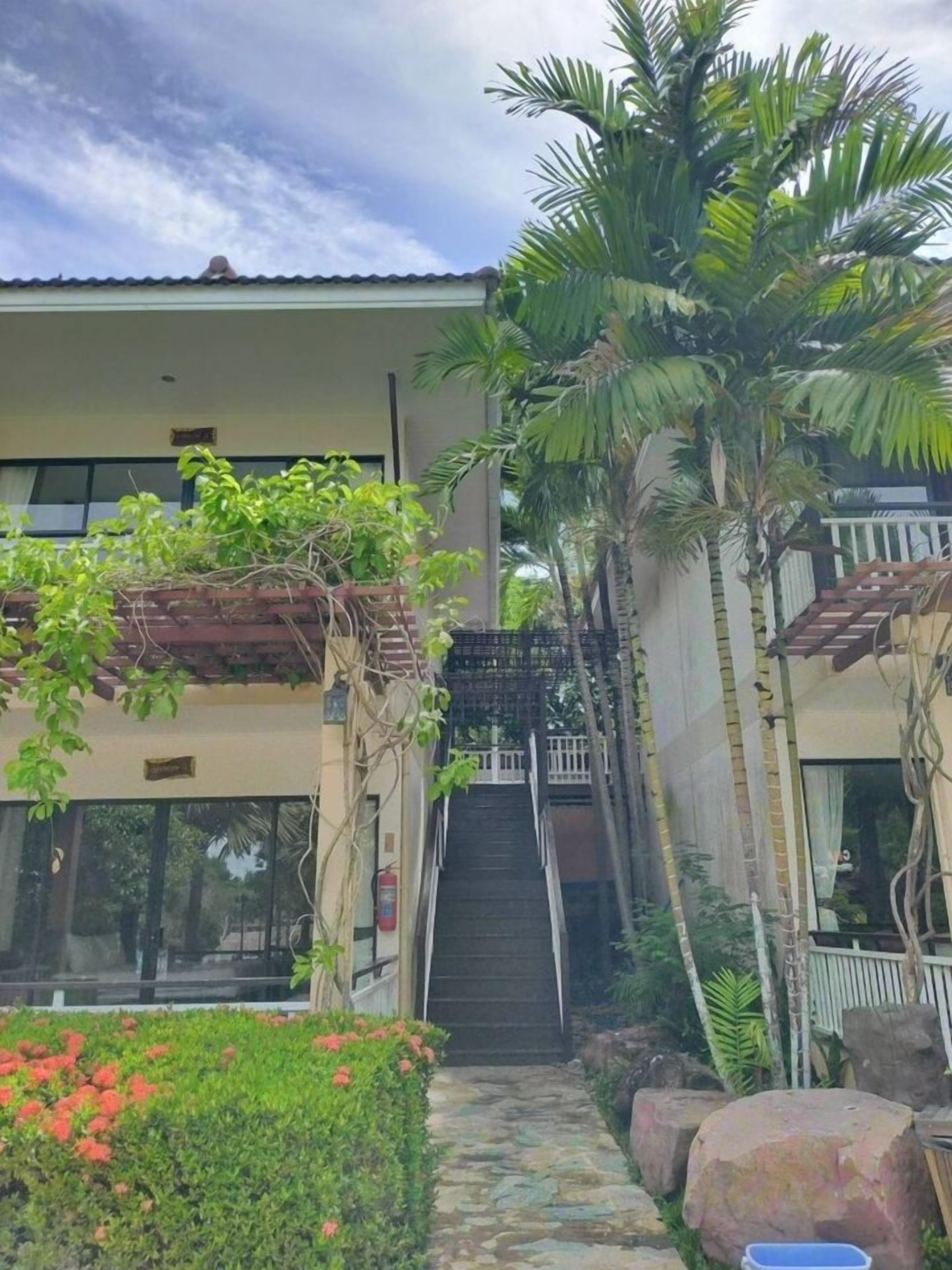 A building with a staircase leading to an upper level, surrounded by greenery and a stone pathway.