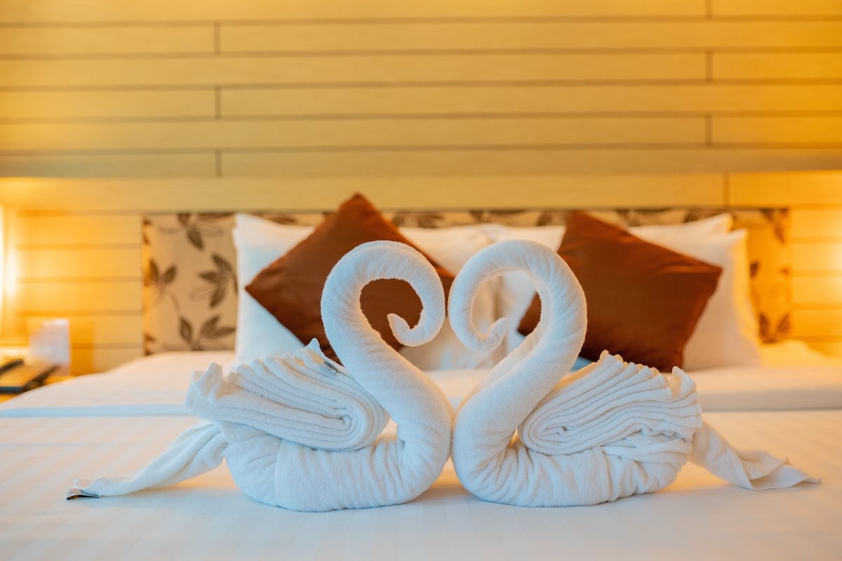 Una cama con dos toallas blancas dispuestas en forma de cisnes, un cabecero de madera y almohadas estampadas.