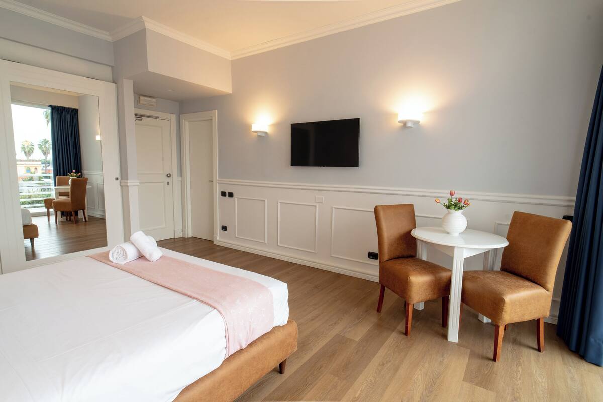 A hotel room with a bed, a small table with two chairs, a TV mounted on the wall, and a view of the outside balcony.