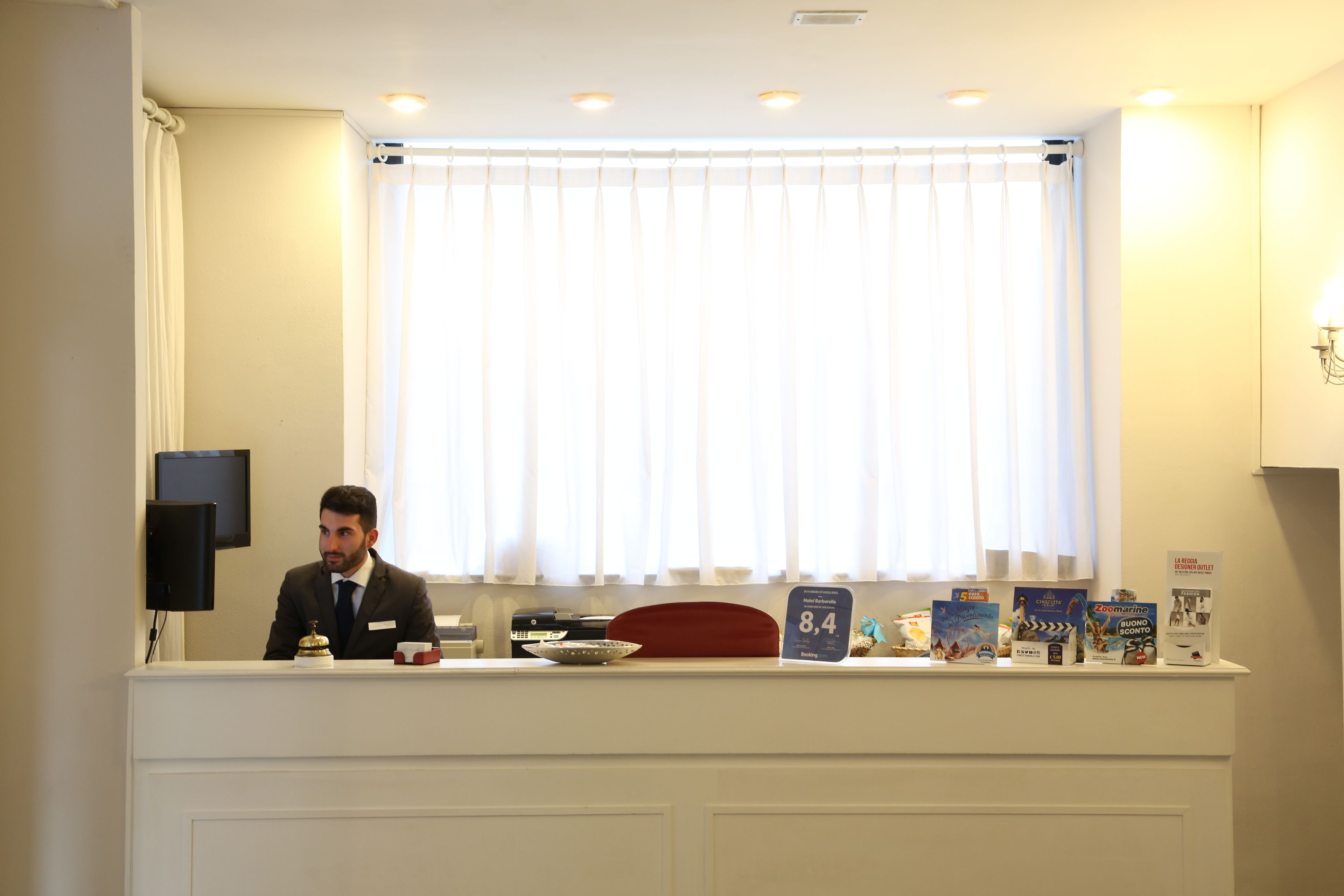Un mostrador de recepción de hotel con un hombre sentado detrás, una ventana con cortinas y varios objetos sobre la encimera, incluyendo un reloj, un cojín rojo y material promocional.