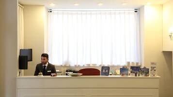 Un mostrador de recepción de hotel con un hombre sentado detrás, una ventana con cortinas y varios objetos sobre la encimera, incluyendo un reloj, un cojín rojo y material promocional.