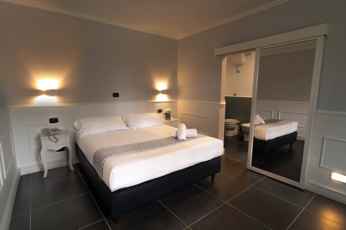 A modern bedroom with a large bed, bedside tables, a mirror, and a bathroom visible through an open door.
