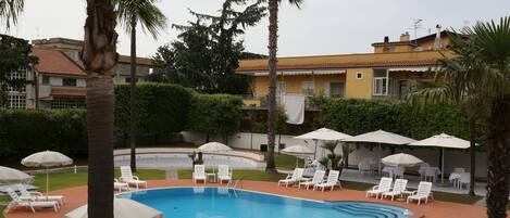 Una piscina con varias secciones, rodeada de sillas de descanso blancas y sombrillas, con un edificio al fondo.