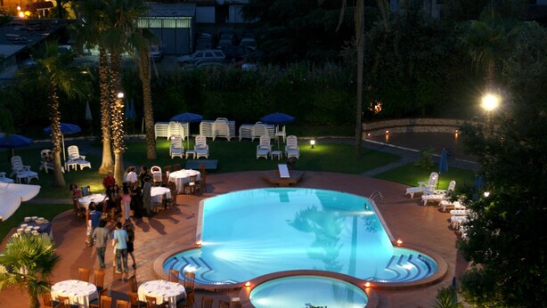 A pool area with lounge chairs, umbrellas, and tables, surrounded by palm trees and greenery. - Hotel Barbarella (Giugliano in Campania)