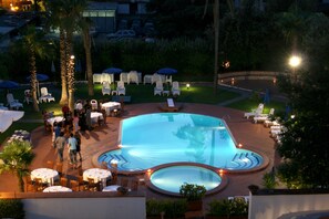 A pool area with lounge chairs, umbrellas, and tables, surrounded by palm trees and greenery. - Hotel Barbarella (Giugliano in Campania)