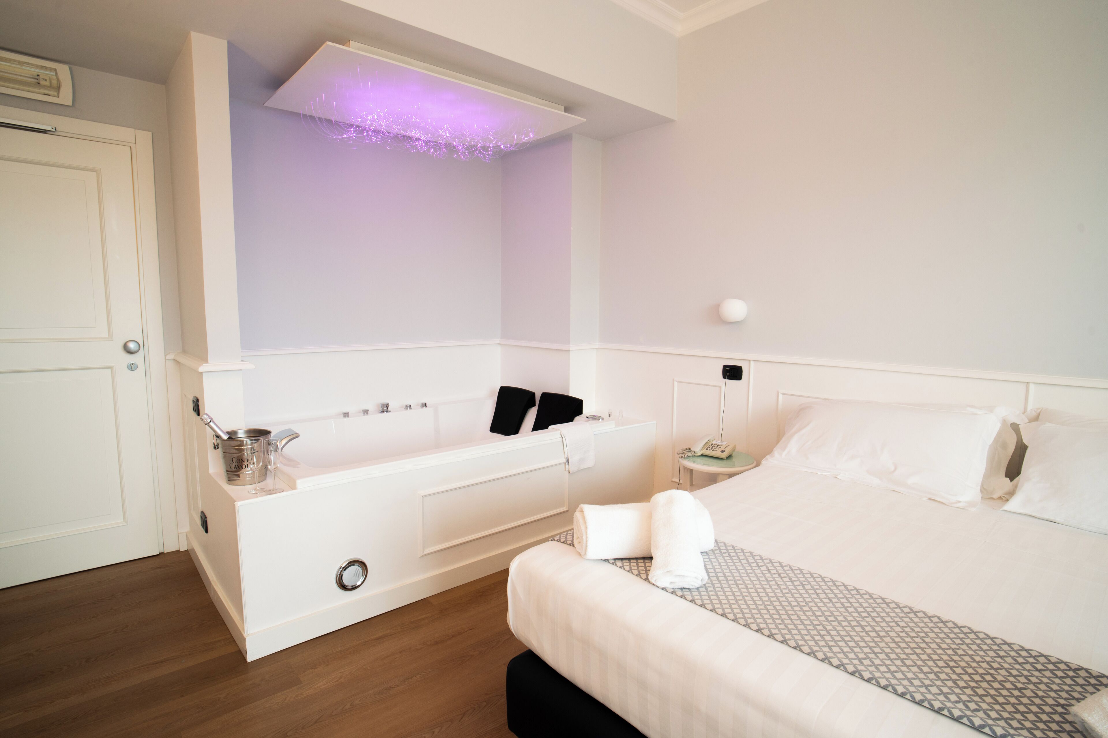 A modern hotel room with a built-in bathtub, a bedside table with a lamp, and a neatly made bed.