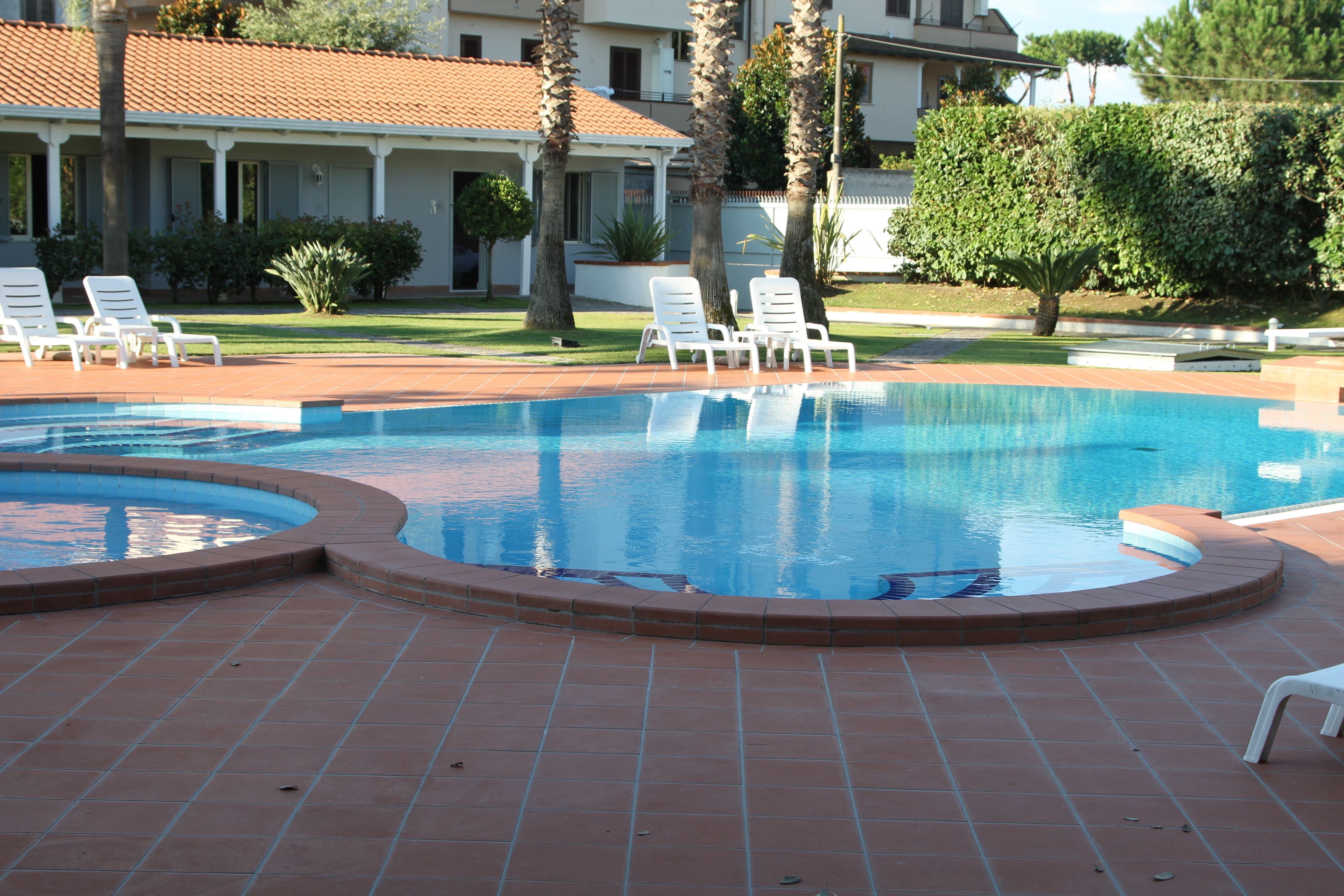 A swimming pool with a tiled deck, surrounded by white lounge chairs and palm trees.