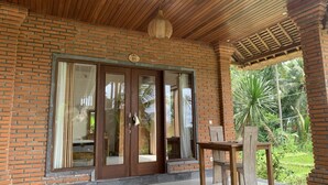 A covered patio with a brick wall, a sliding glass door, and a wooden table with chairs. - Bagiya Suites & Villas (Ubud)