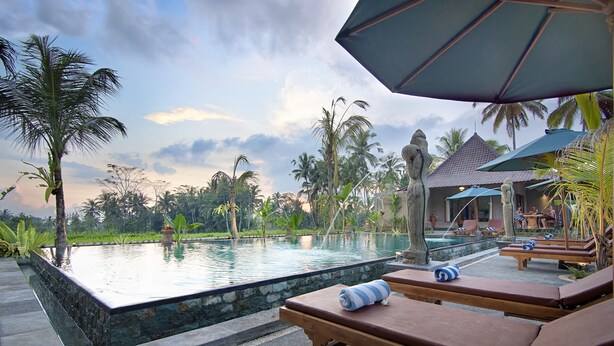 A poolside area with lounge chairs, a statue, and a view of palm trees and a building in the background. - Bagiya Suites & Villas (Ubud)