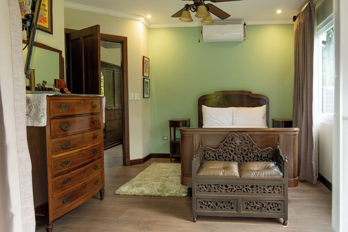 A bedroom with a wooden dresser, a bed with a headboard, a ceiling fan, and a window with curtains.