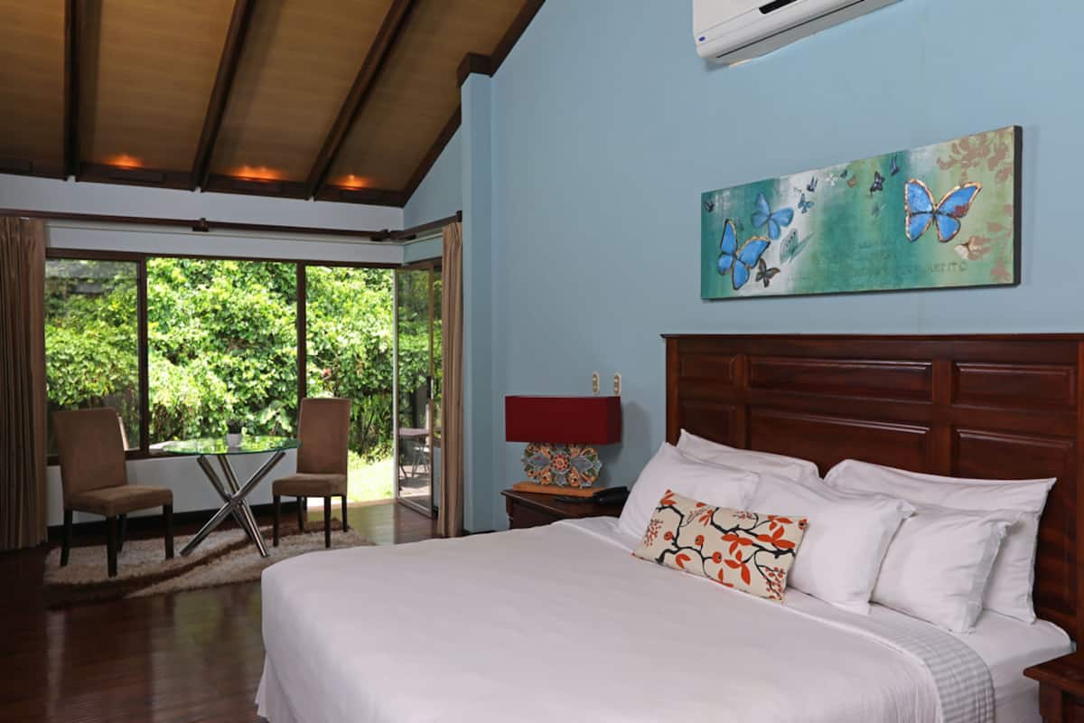 A bedroom with a large bed, a wooden headboard, a painting of butterflies, a nightstand with a lamp, and a view of greenery through the window.