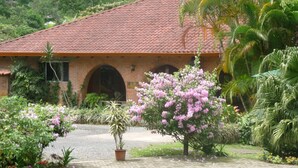 Ein Haus mit rotem Ziegeldach, Backsteinwänden und einem Garten mit rosa Blumen und Palmen.