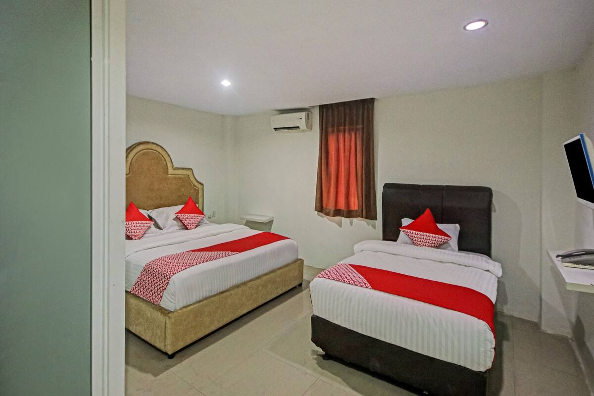 A hotel room with two beds, each with red and white bedding, a wooden headboard, a bedside table, a TV, and an air conditioning unit.