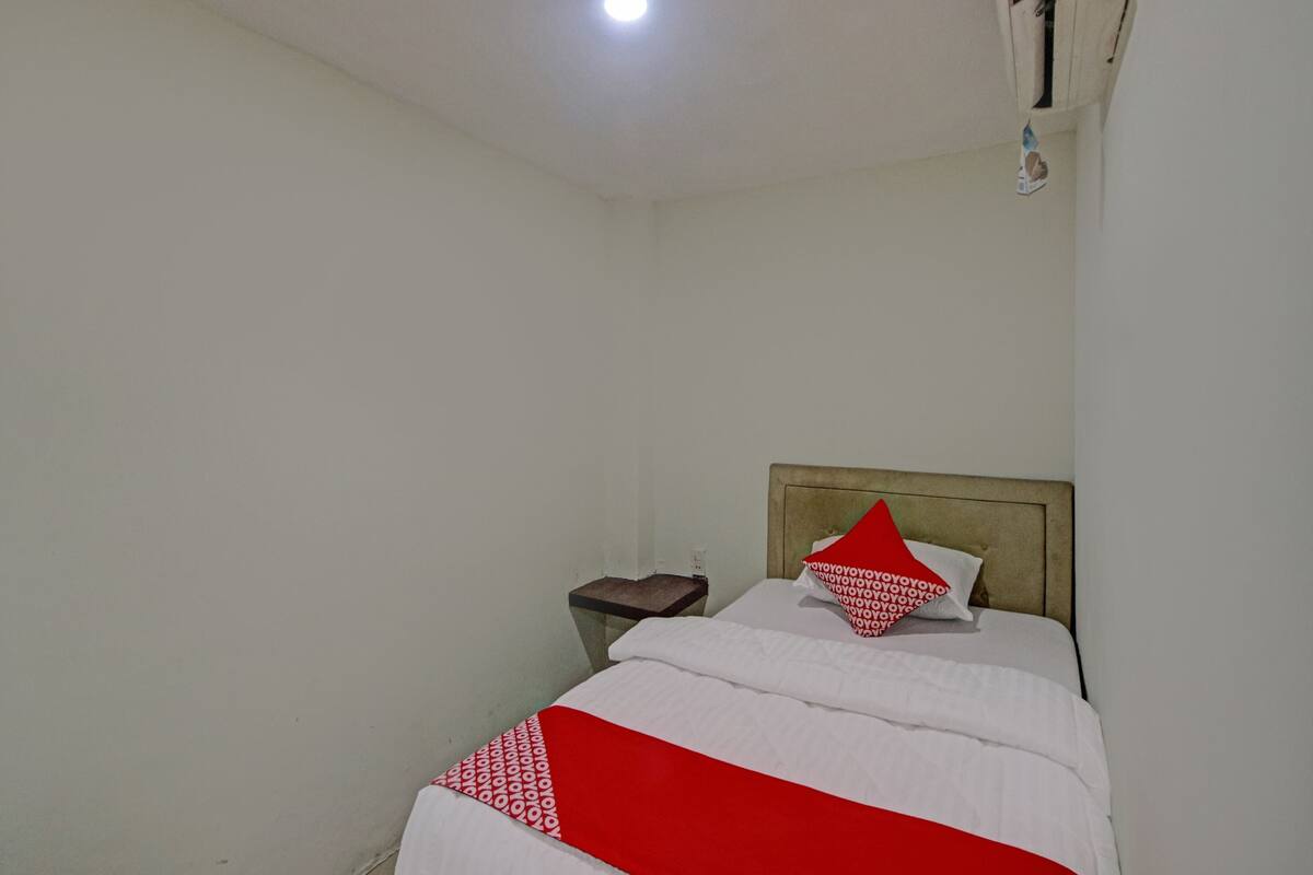 A small, sparsely furnished room with a single bed, a nightstand, and a wall-mounted shelf.
