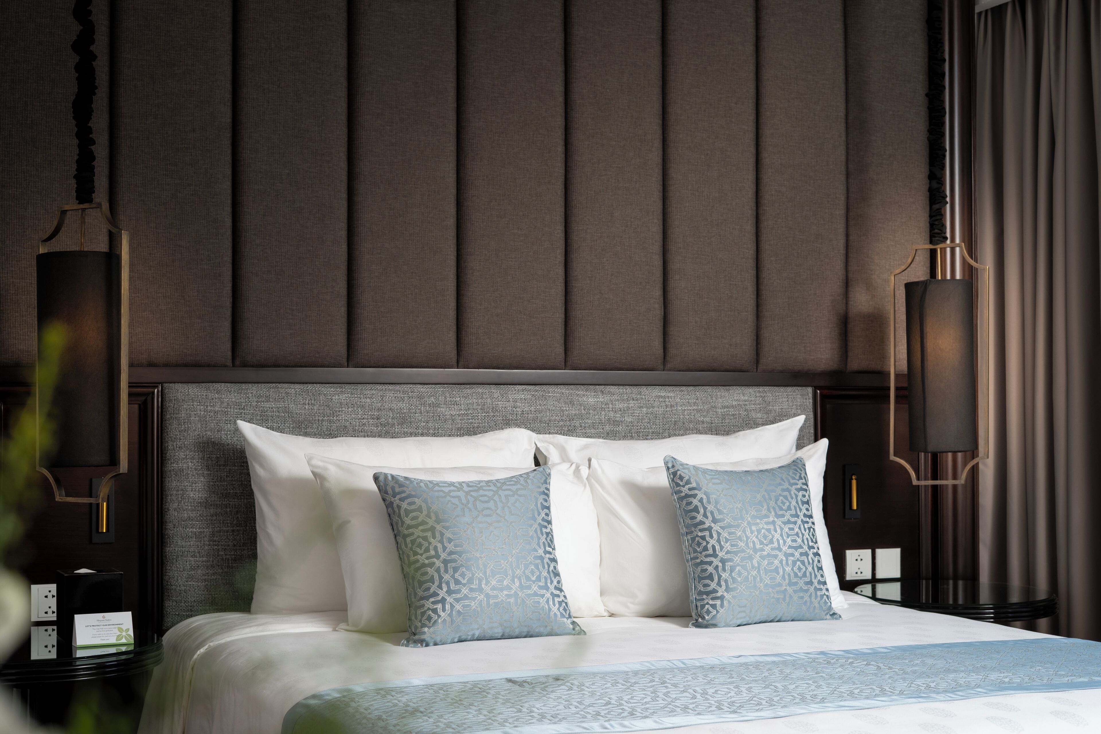 Una cama bien tendida con almohadillas blancas y azules estampadas, un cabecero tapizado en gris y dos lámparas fijadas a la pared.