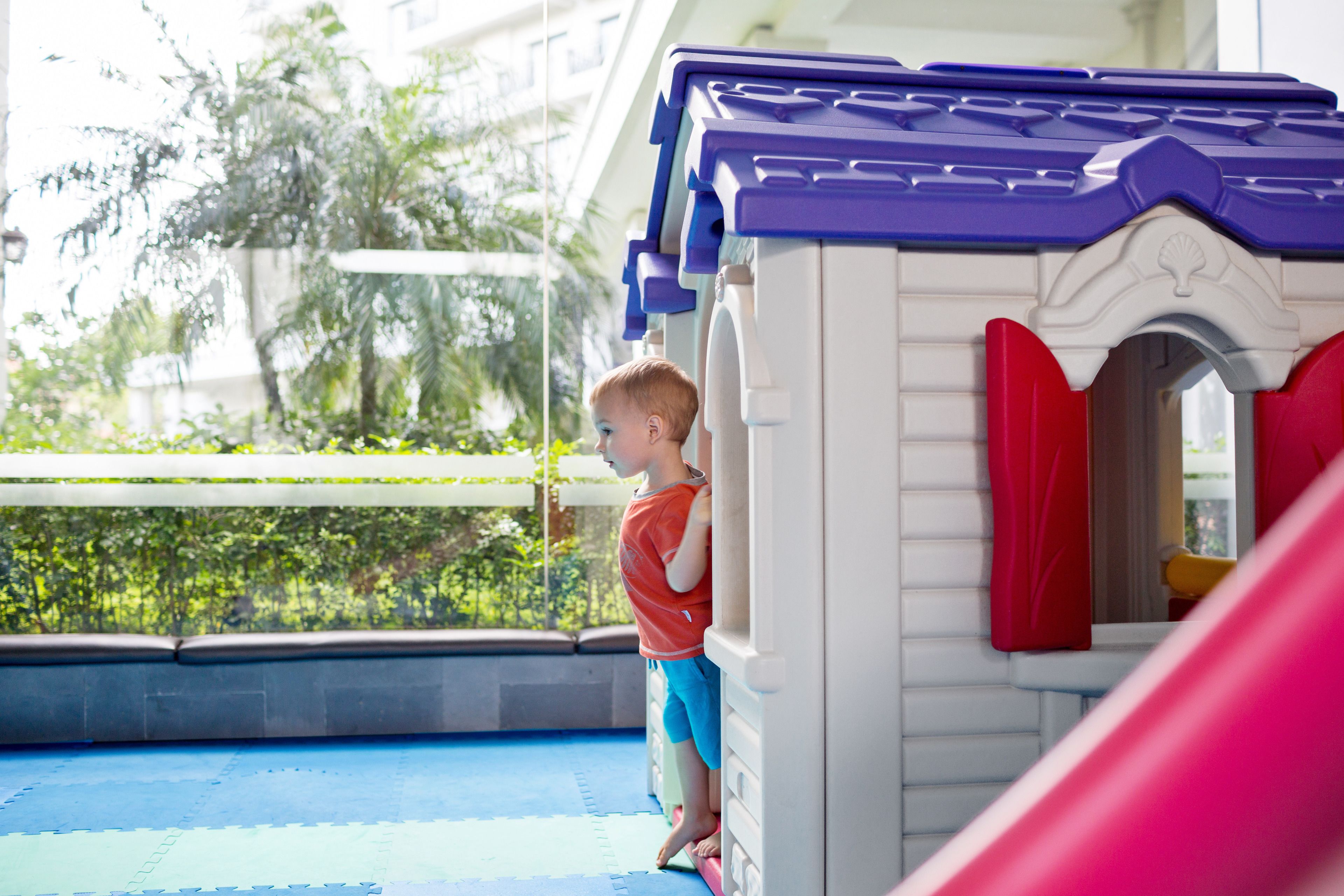 A child standing at the entrance of a playhouse with a blue roof and red shutters.
