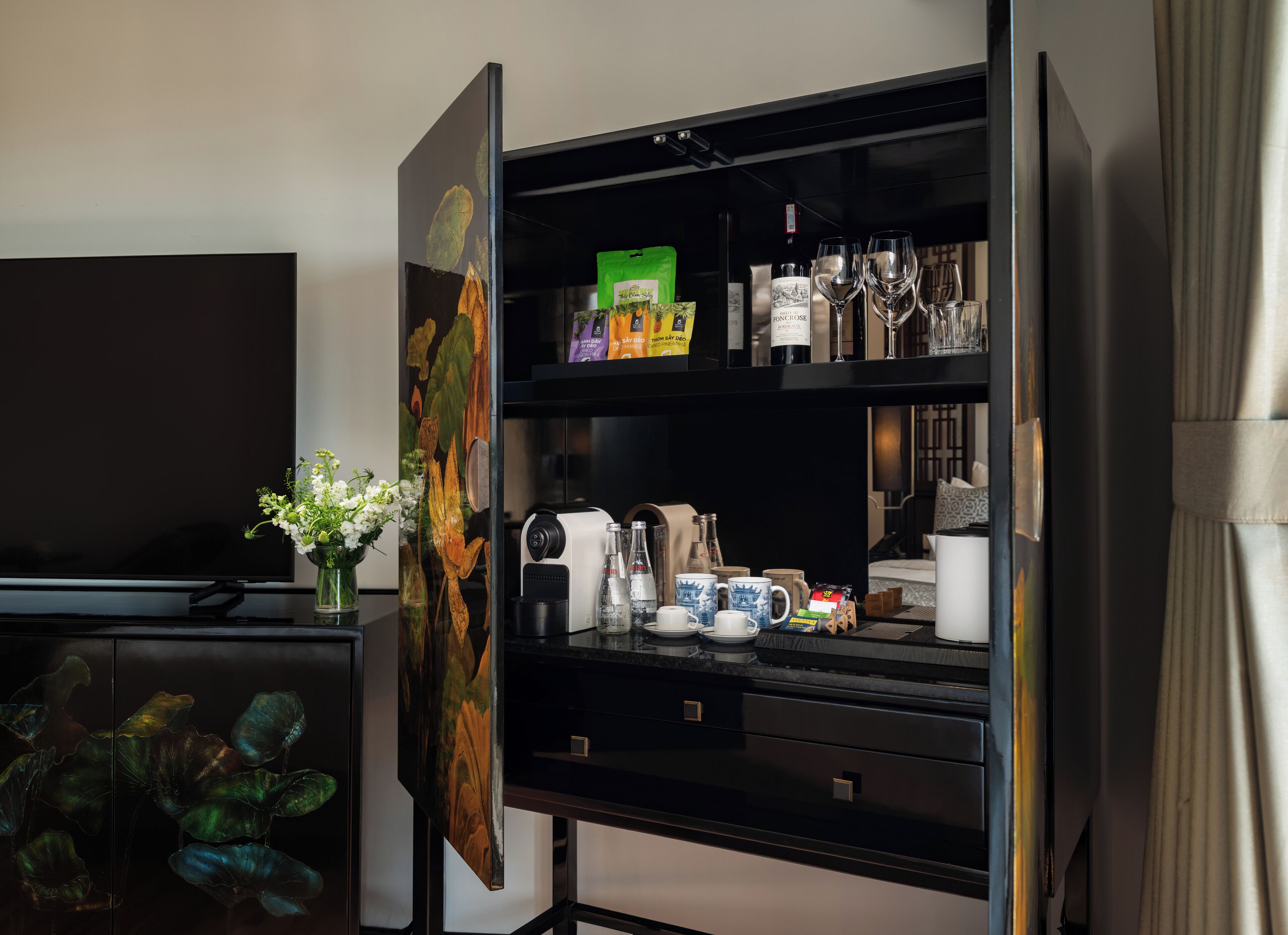 Una habitación con un mueble negro de entretenimiento que contiene un televisor, un cuadro y diversos objetos como una cafetera, tazas y una botella de vino.