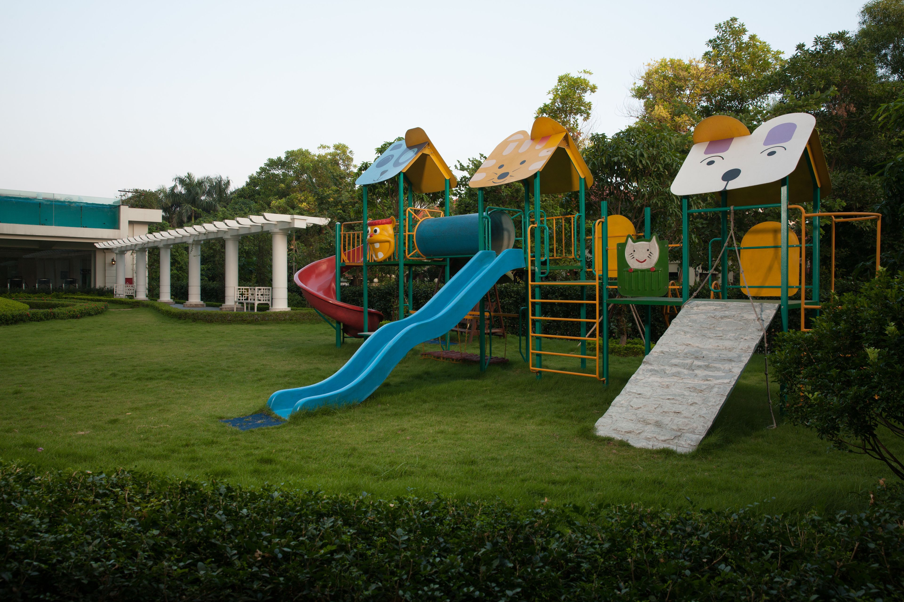 A playground with slides and climbing structures, surrounded by greenery and a building in the background.