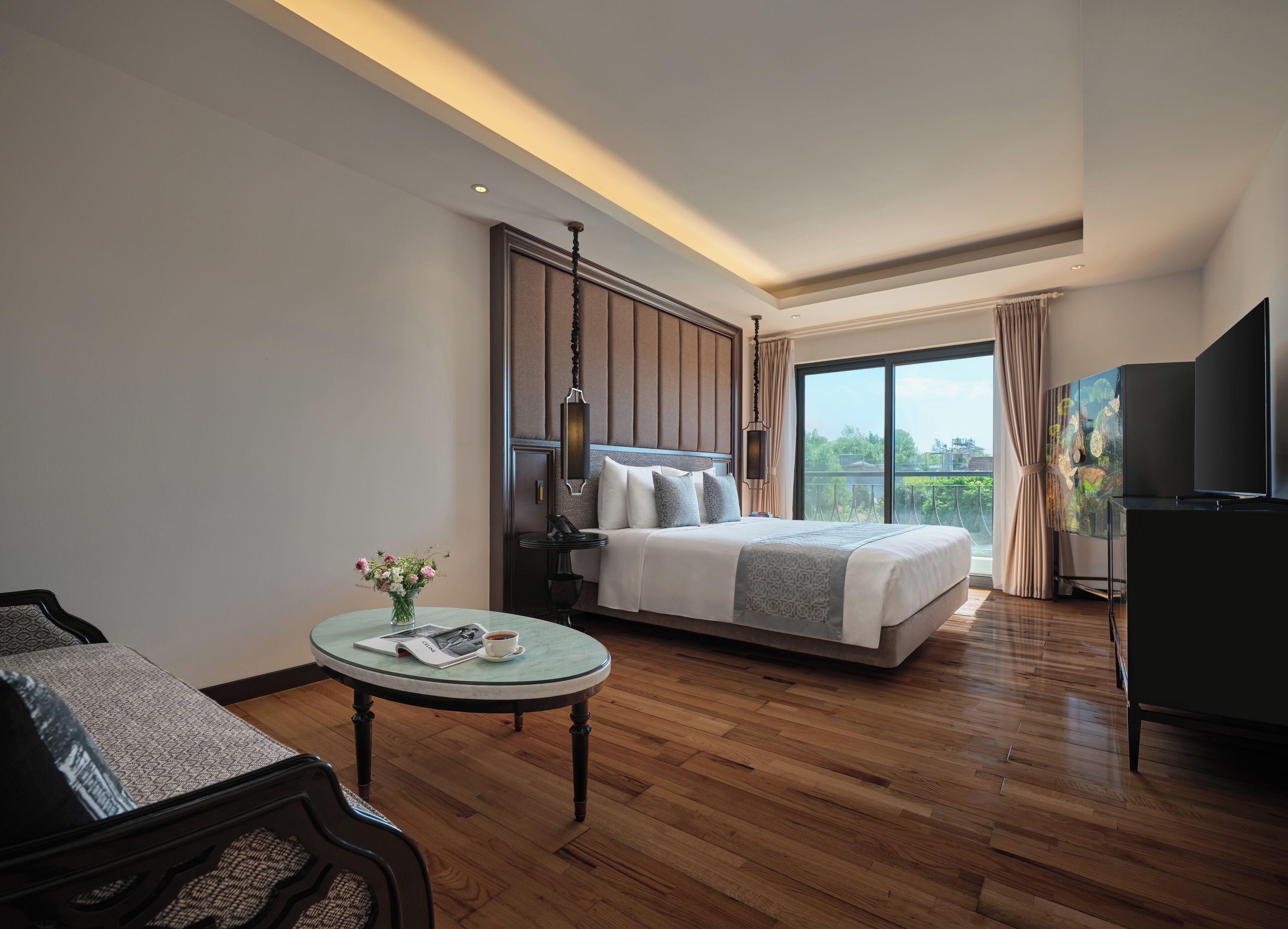 A modern hotel room with a large bed, a small round table with a vase of flowers, a flat-screen TV, and a balcony with a view.