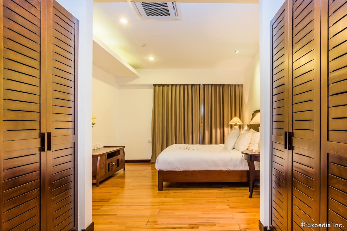 A hotel room with a wooden floor, a bed with white linens, a nightstand with a lamp, and a window with curtains.