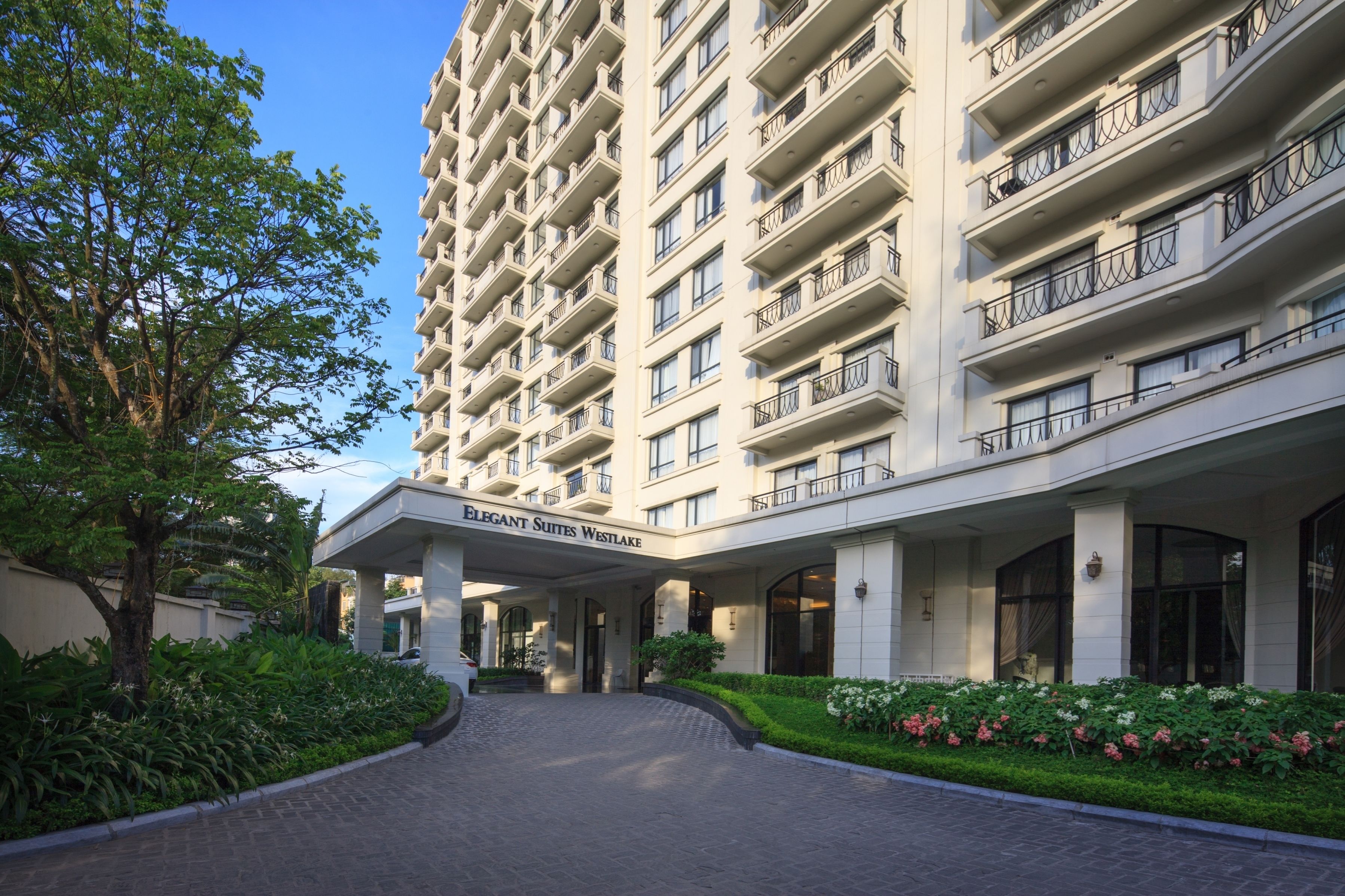 A multi-story hotel with a central entrance, surrounded by lush greenery and a paved driveway.