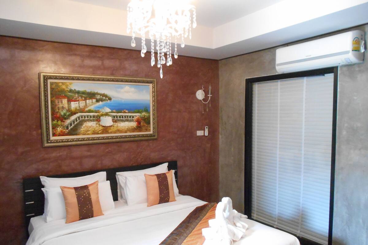 A hotel room with a large bed, a painting on the wall, a chandelier, and an air conditioning unit.
