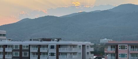A multi-story residential building with balconies, set against a backdrop of mountains and a sunset.