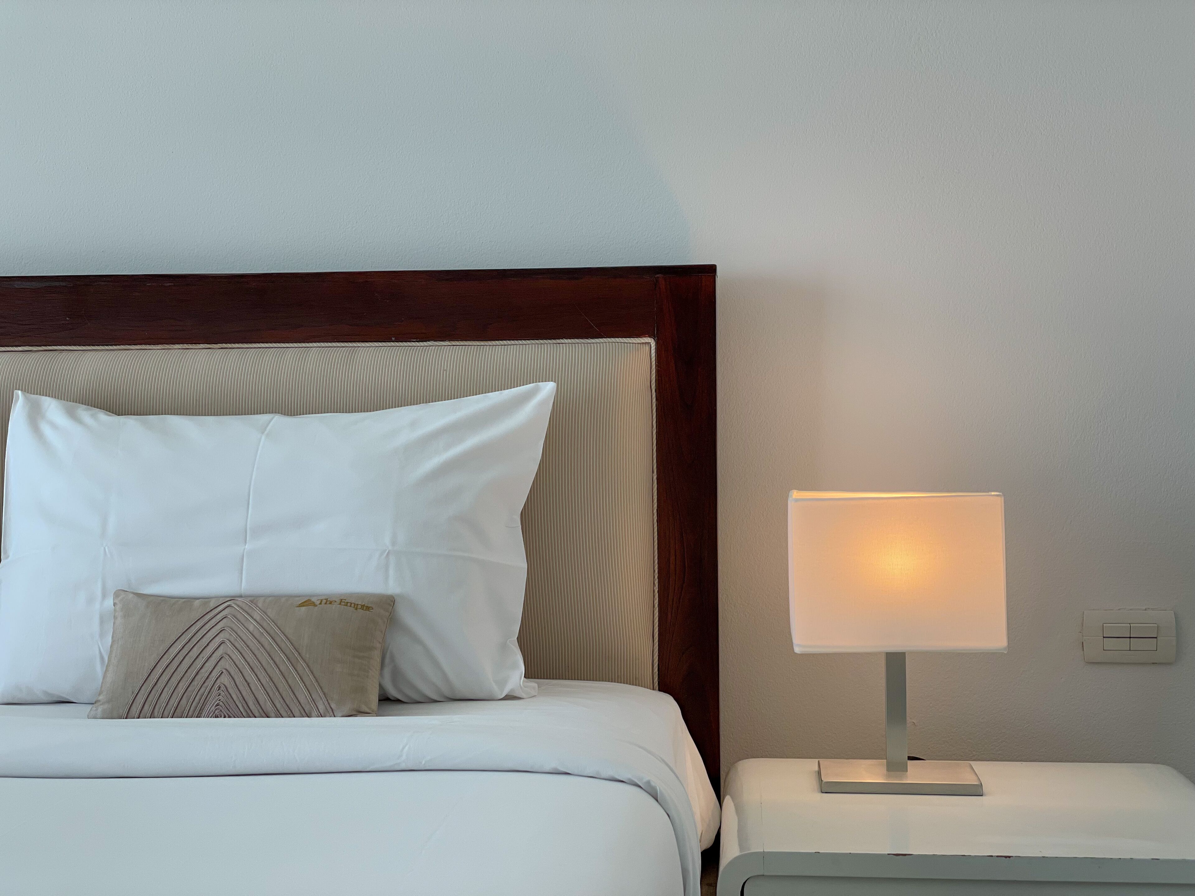 A neatly made bed with white bedding and a single decorative pillow, a bedside table with a lamp, and a wooden headboard.
