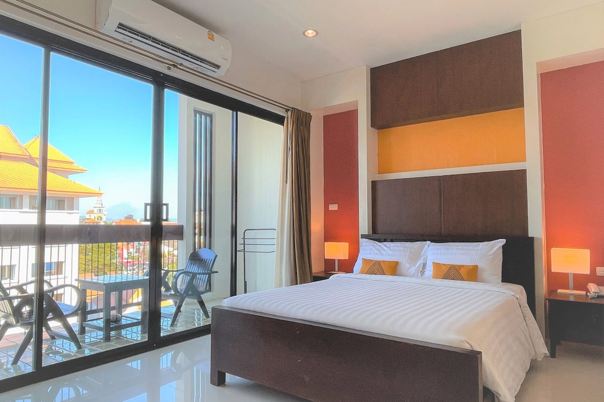 A hotel room with a large bed, two bedside lamps, a view of a building with a yellow roof, and a balcony with chairs.