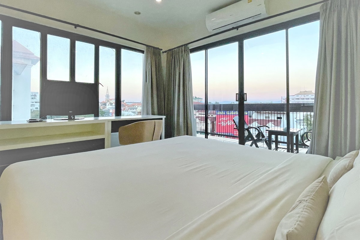 A hotel room with a large bed, a desk with a computer, and a view of a cityscape through large windows.