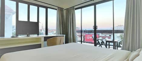 A hotel room with a large bed, a desk with a computer, and a view of a cityscape through large windows.